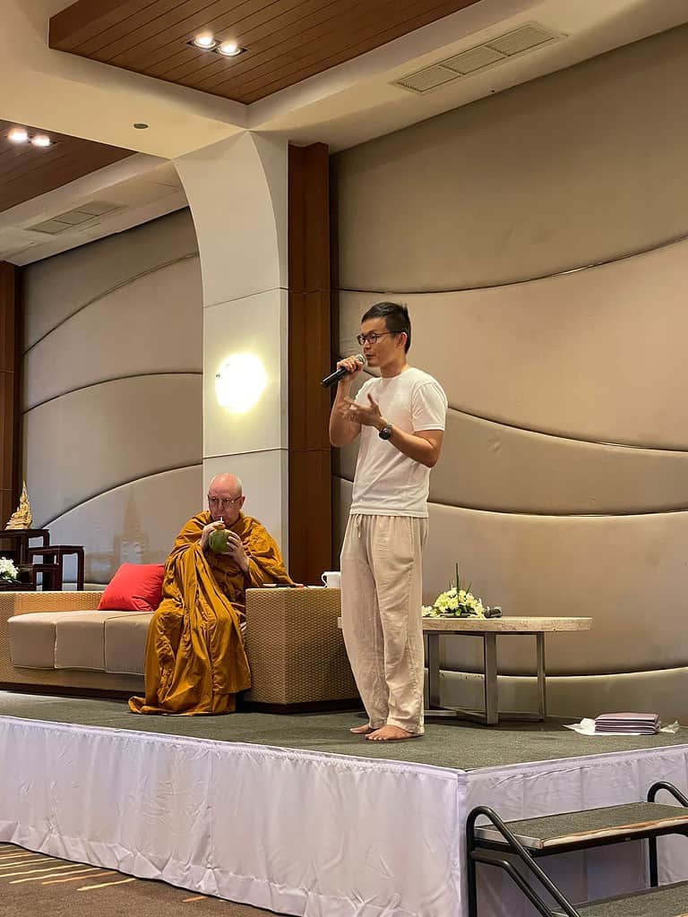 Man holding microphone on a stage, next to a monk on a sofa drinking from a coconut.